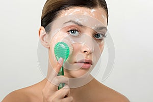 Beautiful young girl cleansing face with cosmetic facial skincare foam, gel or soap by brush