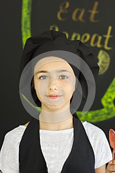 Beautiful young girl cook chef in a black chef`s cap looks direc