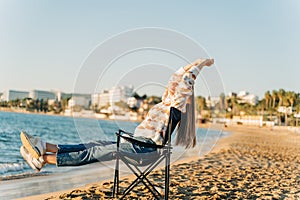 Beautiful young girl in cozy sweater stretching while sitting on foldable chair on winter beach sand. Woman enjoying