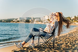 Beautiful young girl in cozy sweater and sunglasses stretching while sitting on foldable chair on winter beach sand