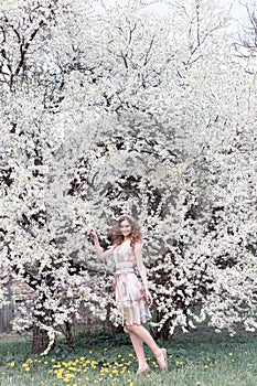 Beautiful young girl with curls in an air colored dress with a wreath on her head walking in the park near a cherry blossom