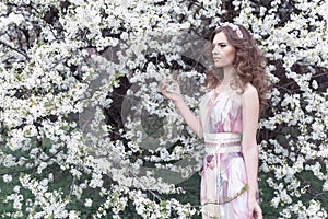 Beautiful young girl with curls in an air colored dress with a wreath on her head walking in the park near a cherry blossom