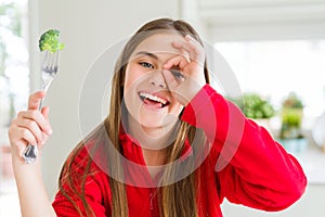 Beautiful young girl eating fresh broccoli with happy face smiling doing ok sign with hand on eye looking through fingers