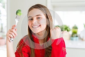 Beautiful young girl eating fresh broccoli pointing and showing with thumb up to the side with happy face smiling