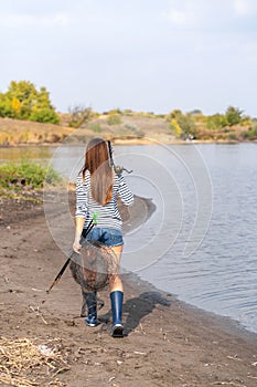 A beautiful young girl goes fishing. A girl with a fishing rod and a cage in hand is walking along the lake