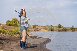 A beautiful young girl goes fishing. A girl with a fishing rod and a cage in hand is walking along the lake