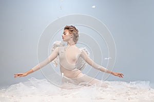 Beautiful young girl, graceful ballerina in image of angel with wings sitting on cloud  on white gray studio