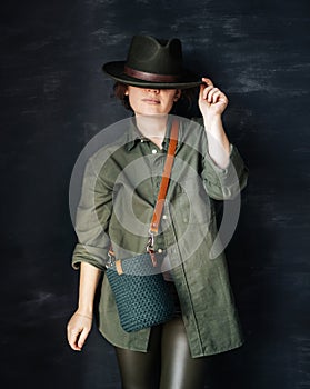 Beautiful young girl in a green shirt and hat holds a knitted bag on a handmade leather belt on a dark