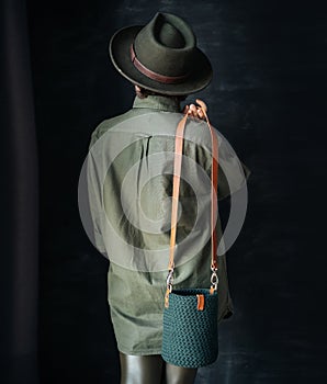 Beautiful young girl in a green shirt and hat holds a knitted bag on a handmade leather belt on a dark background stands with her