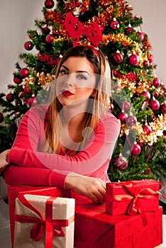 Beautiful young girl with hand on Christmas gift box and reindeer horn on head and Xmas tree