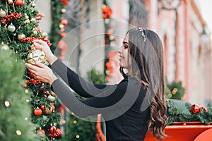 Beautiful young girl hangs balls on a Christmas tree near the store. Cute smiling girl decorates a Christmas tree