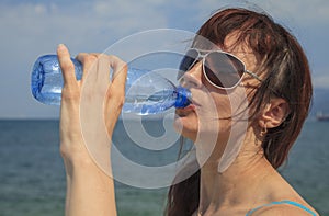 Beautiful young girl having fun drinking water from a plastic bottle, in hot weather near the sea