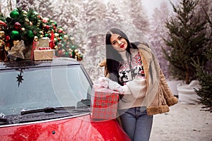 Beautiful young girl is holding gift box near red car with decorated xmas tree on the roof, holiday and new years