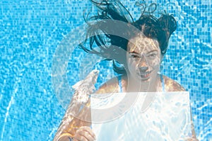 Beautiful young girl holding white blank board in swimming pool under water, family vacation