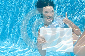 Beautiful young girl holding white blank board in swimming pool under water, fun on family vacation