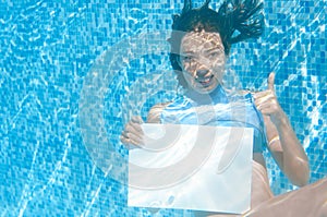 Beautiful young girl holding white blank board in swimming pool under water, fun on family vacation