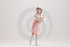 Beautiful young girl in image of retro cater waiter wearing 70s, 80s fashion style uniform isolated over light studio