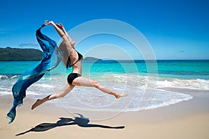 Beautiful young girl jumping with waving blue cape, scarf, on tropical beach