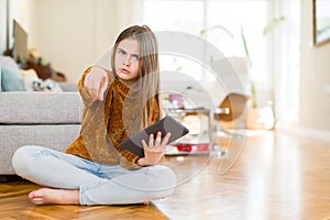 Beautiful young girl kid using digital touchpad tablet sitting on the floor pointing with finger to the camera and to you, hand