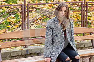 Beautiful young girl with large autumn sad eyes in a coat and ripped black jeans sitting on a bench in autumn park