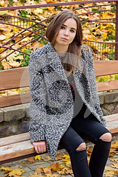 Beautiful young girl with large autumn sad eyes in a coat and ripped black jeans sitting on a bench in autumn park