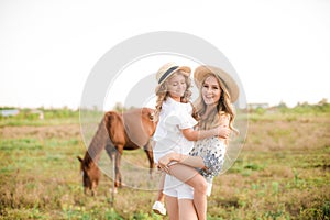 A beautiful young girl, with light curly hair in a straw hat with a little sister hugging and laughing near horses