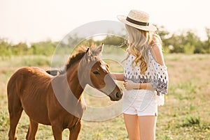 A beautiful young girl, with light curly hair in a straw hat stroking the foal`s nose in the countryside, warm autumn