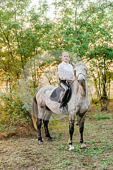 Beautiful young girl with light hair in uniform competition smiling and astride a horse in sunset