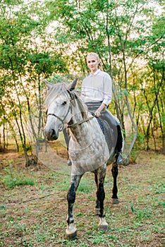 Beautiful young girl with light hair in uniform competition smiling and astride a horse in sunset