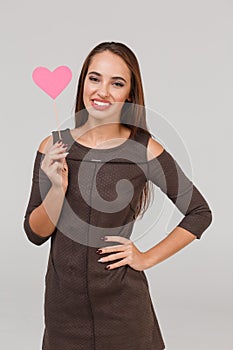 A young girl with long dark hair is holding a pink heart on a stick and smiling. Isolation.