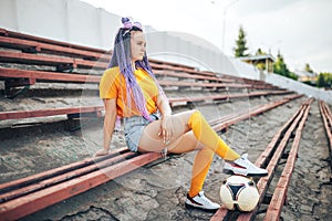 Beautiful young girl with long hair and a soccer ball sitting in a stadium, unusual hairstyle
