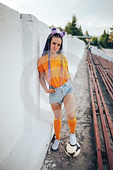 Beautiful young girl with long hair and a soccer ball sitting in a stadium, unusual hairstyle