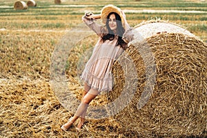 Beautiful young girl with long hair in sunnglasses and straw hat posing on a wheat field near hay bales. Happy brunette in summer