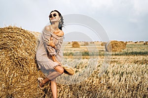 Beautiful young girl with long hair in sunnglasses and straw hat posing on a wheat field near hay bales. Happy brunette in summer