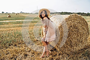 Beautiful young girl with long hair in sunnglasses and straw hat posing on a wheat field near hay bales. Happy brunette in summer