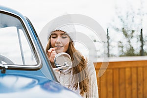 A beautiful young girl looks in the car mirror and paints her lips. Woman with long dark thick hair next to blue retro car in