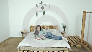 Beautiful young girl lying on the bed reading a glossy magazine