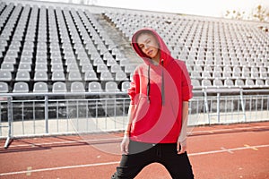 A beautiful young girl of mixed race stands on a sports stadium running track dressed in a red hoodie with a hood.