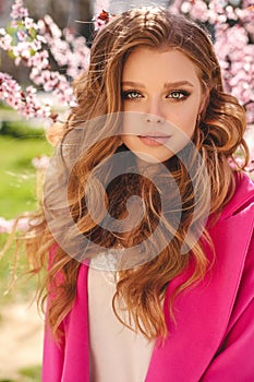 Beautiful young girl with natural hair color in elegant clothes posing among flowering peach trees