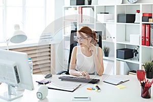 Beautiful young girl in the office working with documents, holding a pen in her hand and looking at the monitor.