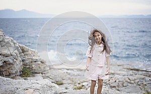 Beautiful young girl in a pink suit poses on a rock high above the sea during sunset. Dark clouds all over the sky. Wind in the