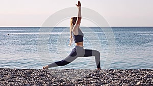 Beautiful young girl practicing yoga, doing warrior pose, stretching on the beach at the seaside