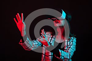 Beautiful young girl reaching out with her hands and wearing virtual reality goggles in studio with color gels lighting