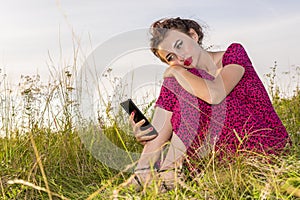 Beautiful young girl in red dress with mobile phone in her hand sits on green field.