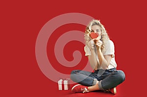 Beautiful young girl with red hearts and a white gift with a bow sits in full growth on an isolated red backgroundValentine`s Day