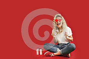 Beautiful young girl with red hearts and a white gift with a bow sits in full growth on an isolated red backgroundValentine`s Day
