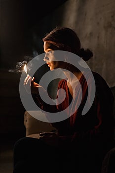 Beautiful young girl in red shirt lights up her pretty face with match sitting in dark room and holding matchbox in hand