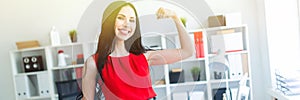 Beautiful young girl in a red suit stands in the office and shows a muscle on her arm.