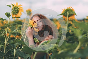 Beautiful young girl with red wavy hair and freckles in stripped colourful dress enjoying nature on the field of sunflowers.