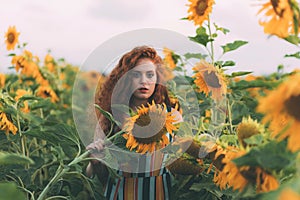 Beautiful young girl with red wavy hair and freckles in stripped colourful dress enjoying nature on the field of sunflowers.